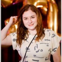 A woman with wavy brown hair wearing a white patterned top stands in front of gold balloons, looking at the camera and smiling.
