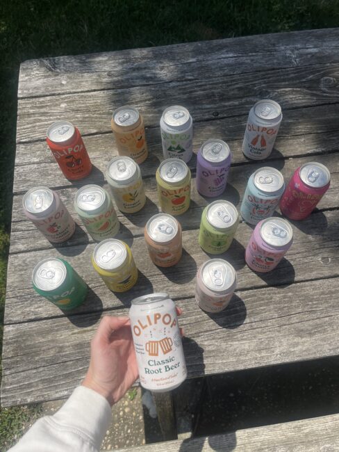 A hand holds an OLIPOP Classic Root Beer can above a wooden table displaying various OLIPOP soda cans arranged in rows on a sunny day.