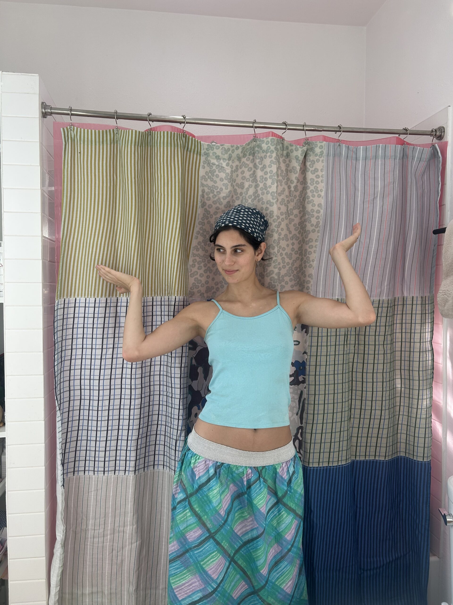 Person in a blue tank top and plaid skirt stands with arms raised in a bathroom, in front of a shower curtain made of various patterned fabrics.
