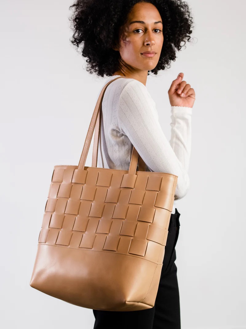 A person wearing a white long-sleeve shirt and black pants holds a large tan woven leather tote bag over one shoulder, standing against a plain light background.