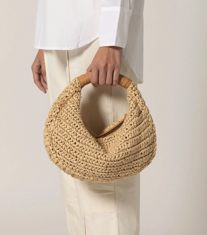 A person wearing white clothes holds a round, woven straw handbag with a wooden handle against a neutral background.