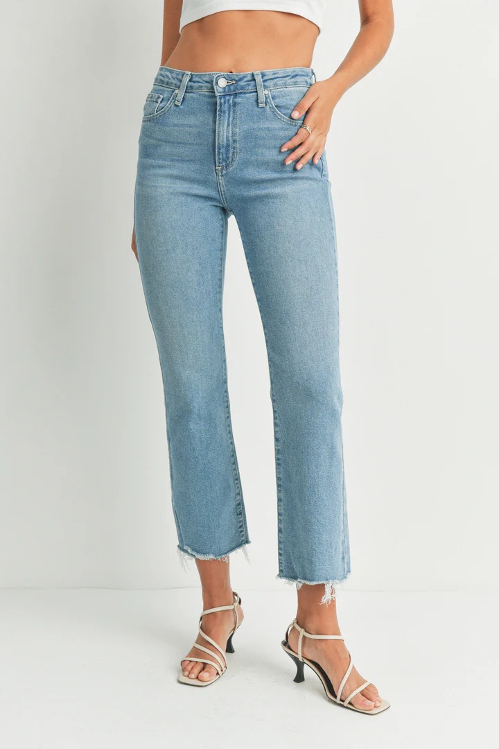 Person wearing light blue high-waisted cropped jeans with raw hems, white strappy heeled sandals, and a white crop top, standing against a plain white background.