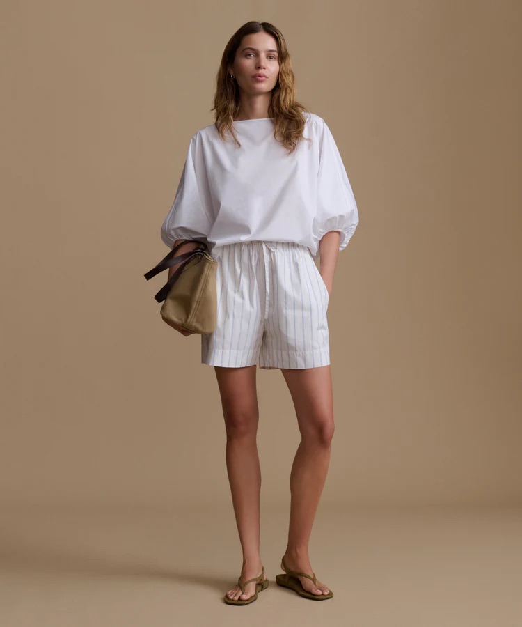 A woman stands against a beige background wearing a white puff-sleeve blouse, striped shorts, brown sandals, and holding a tan clutch.