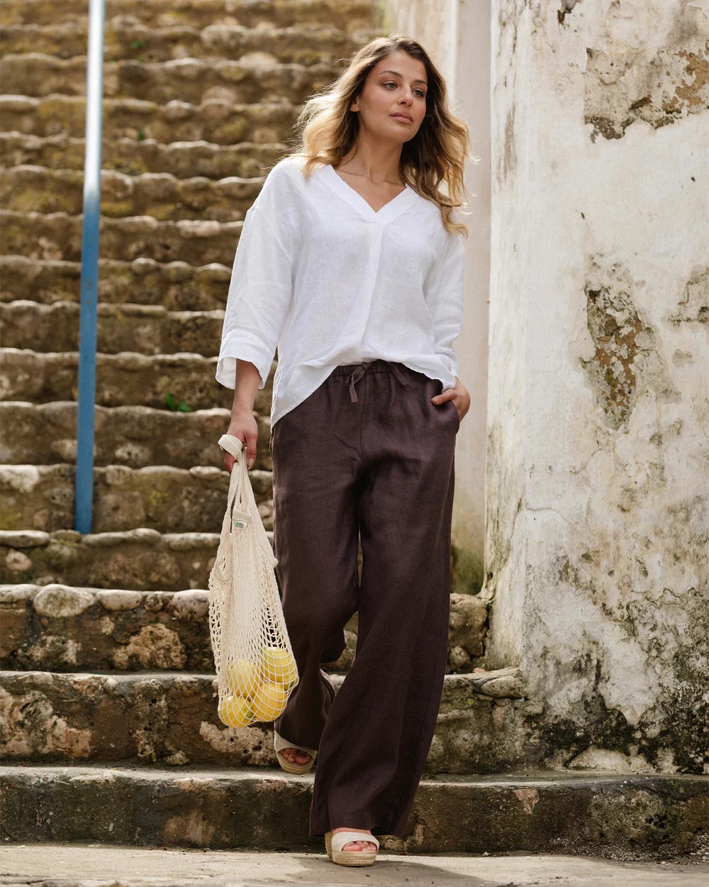 Woman in a white blouse and dark wide-leg pants walks down stone steps, holding a mesh bag with lemons. A weathered wall is beside her.