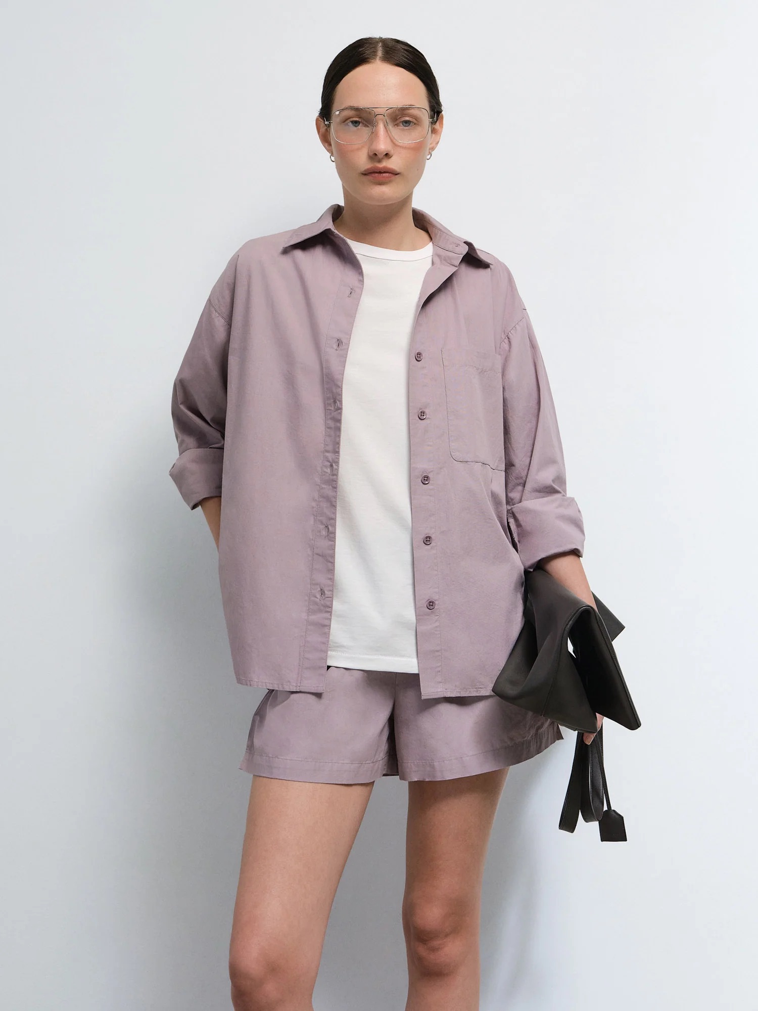 A woman stands against a plain background wearing glasses, a light purple button-up shirt, matching shorts, a white t-shirt, and holding a black clutch bag.