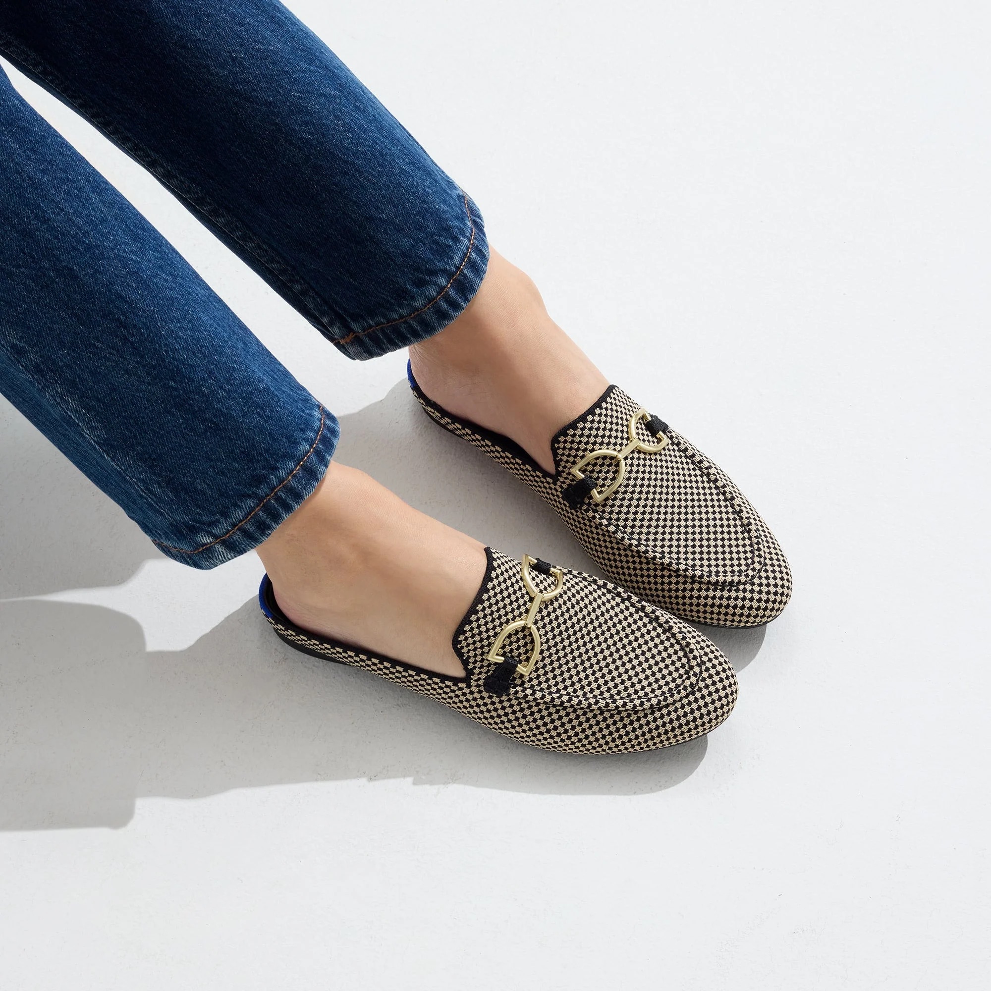 Person wearing blue jeans and houndstooth-patterned slip-on loafers with gold bit detail, standing on a white surface.