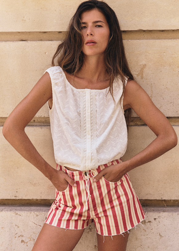 Woman standing against a beige wall, wearing a sleeveless white top and red-and-white vertically striped shorts with hands in pockets.