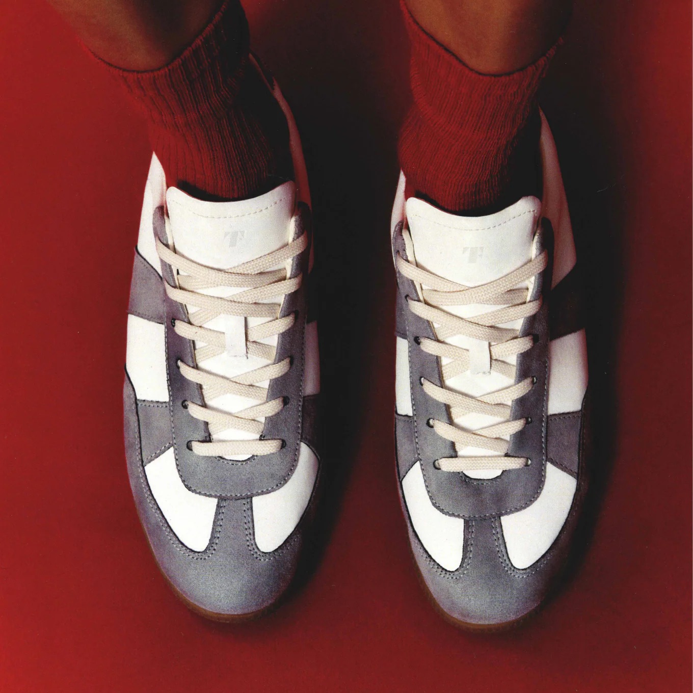 A person wearing white and gray sneakers with beige laces and red socks, standing on a red surface.