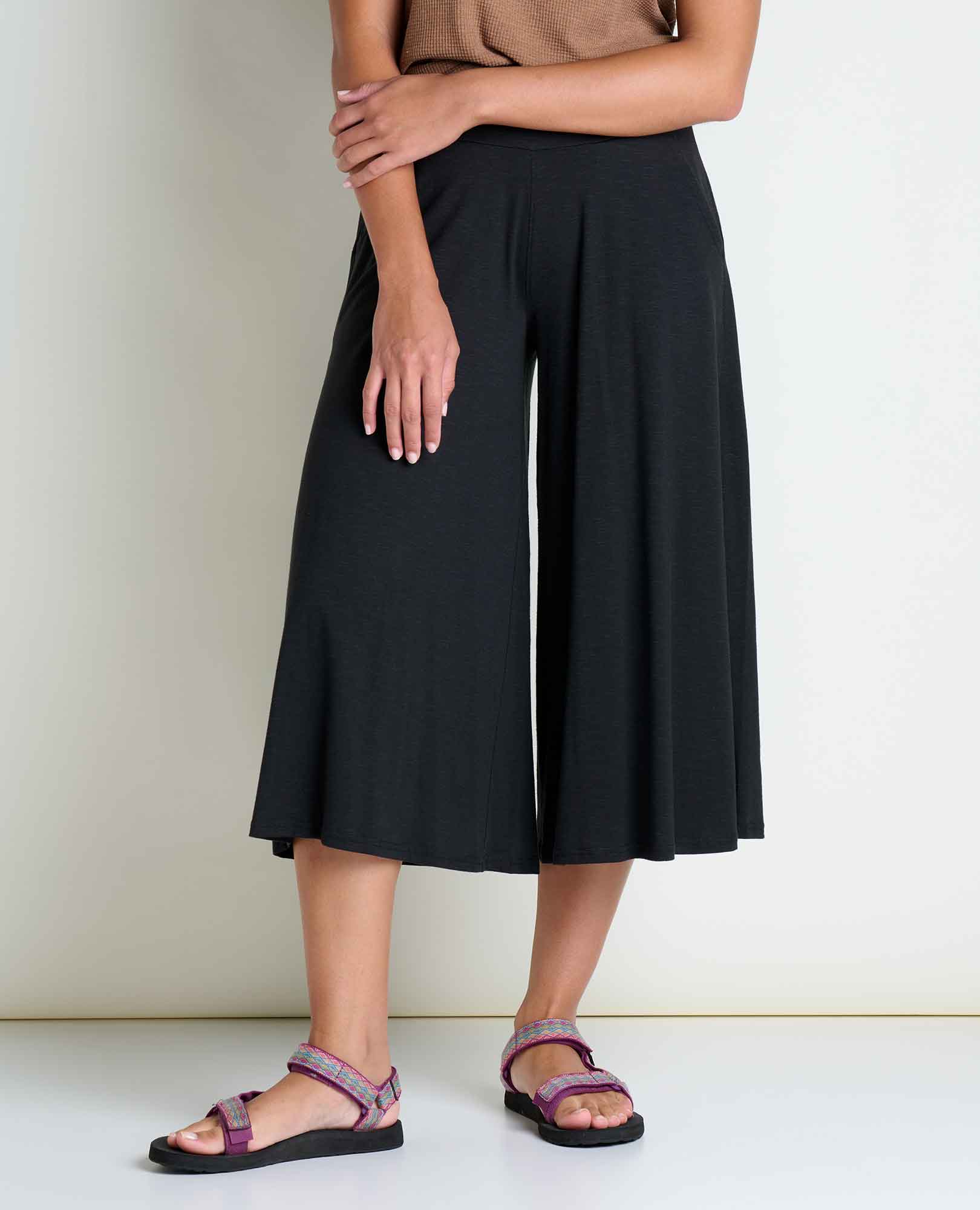 Person wearing black wide-leg culottes, a brown sleeveless top, and purple sandals, standing against a plain light-colored wall with arms crossed over the chest.