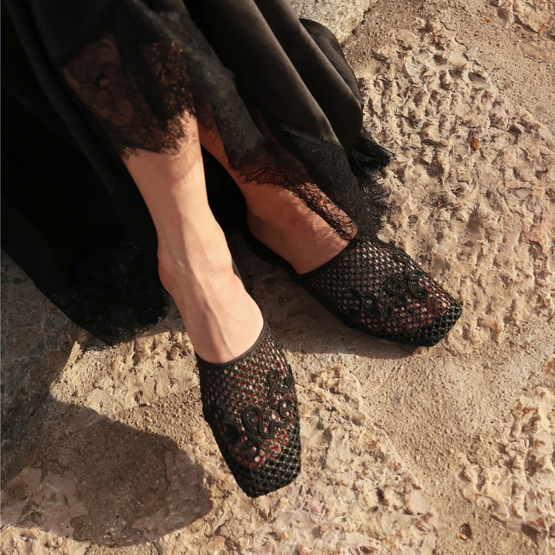 Person wearing a long black lace dress and black mesh slip-on shoes stands on a textured stone surface.