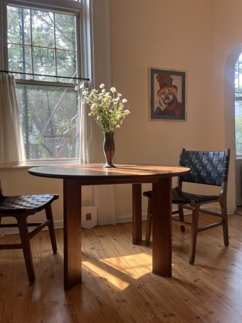 A round wooden table with a vase of daisies sits between two woven chairs in a sunlit room. A framed clown portrait hangs on the wall.