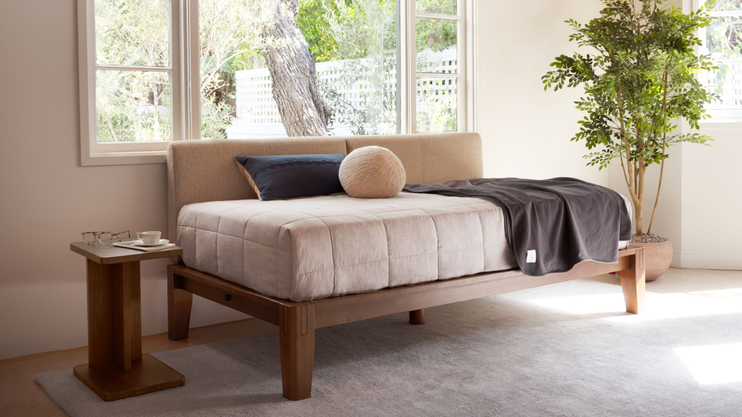 A neatly made bed with beige bedding, dark throw, and two pillows sits by a large window next to a small wooden table with a cup and saucer, in a bright, modern bedroom.