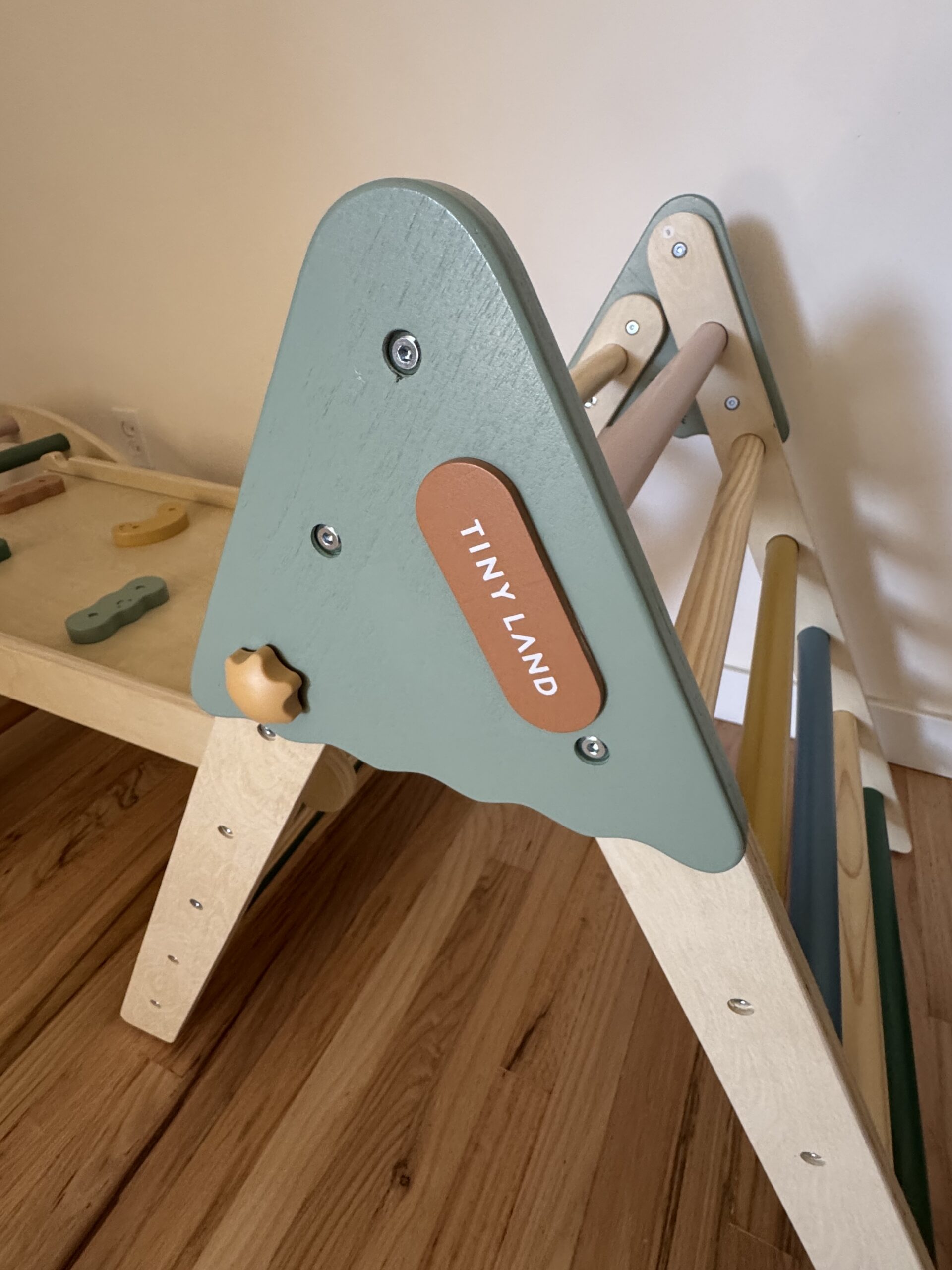 A wooden climbing triangle with green and natural wood tones sits on a hardwood floor. A label reads “TINY LAND.”.