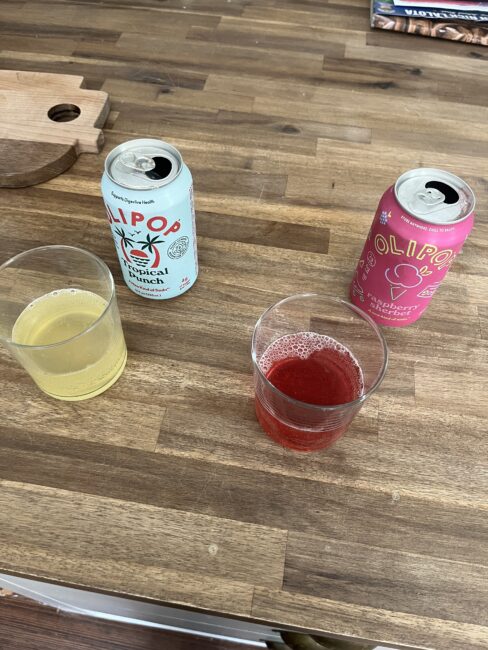 Two cans of Olipop soda, one Tropical Punch and one Raspberry Hibiscus, sit on a wooden table next to glasses filled with matching colored drinks.