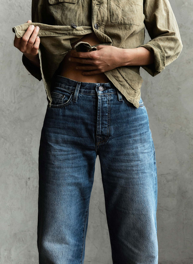 Person wearing a loose olive button-up shirt and high-waisted blue jeans, partially lifting the shirt to reveal their midriff against a plain gray background.