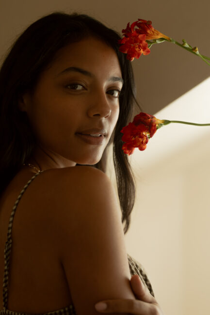 A woman with long dark hair stands indoors, looking at the camera with red flowers near her face and shoulder, in soft natural lighting.