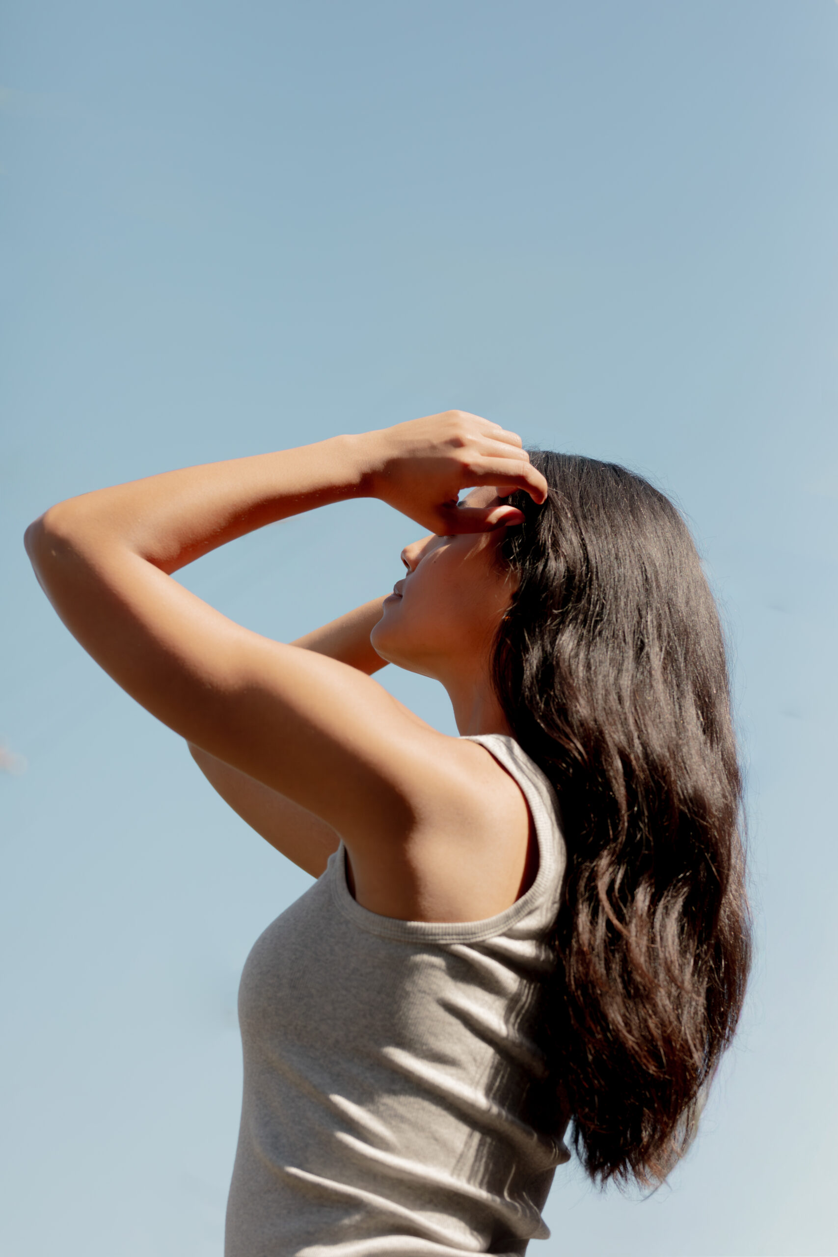 Woman with long dark hair in a sleeveless top stands outdoors, holding her head with both hands and looking up at the clear blue sky.