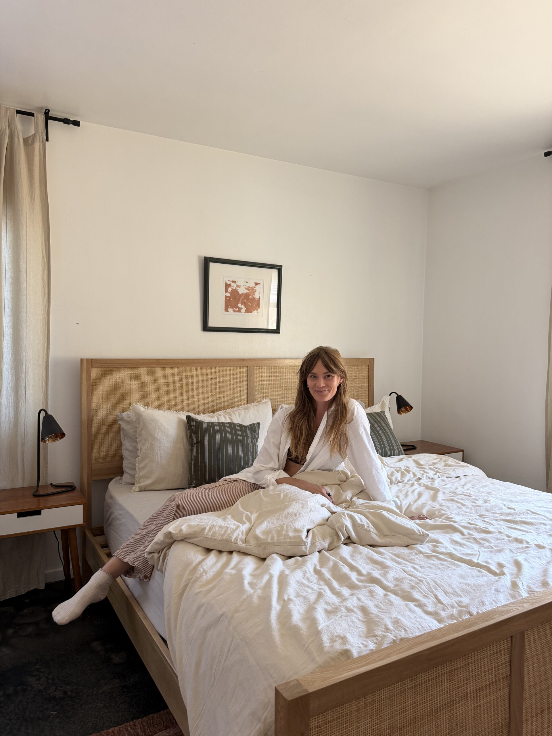 Woman sitting on an unmade bed in a bright, minimal bedroom with neutral decor, wicker headboard, framed art, and two nightstands with lamps.