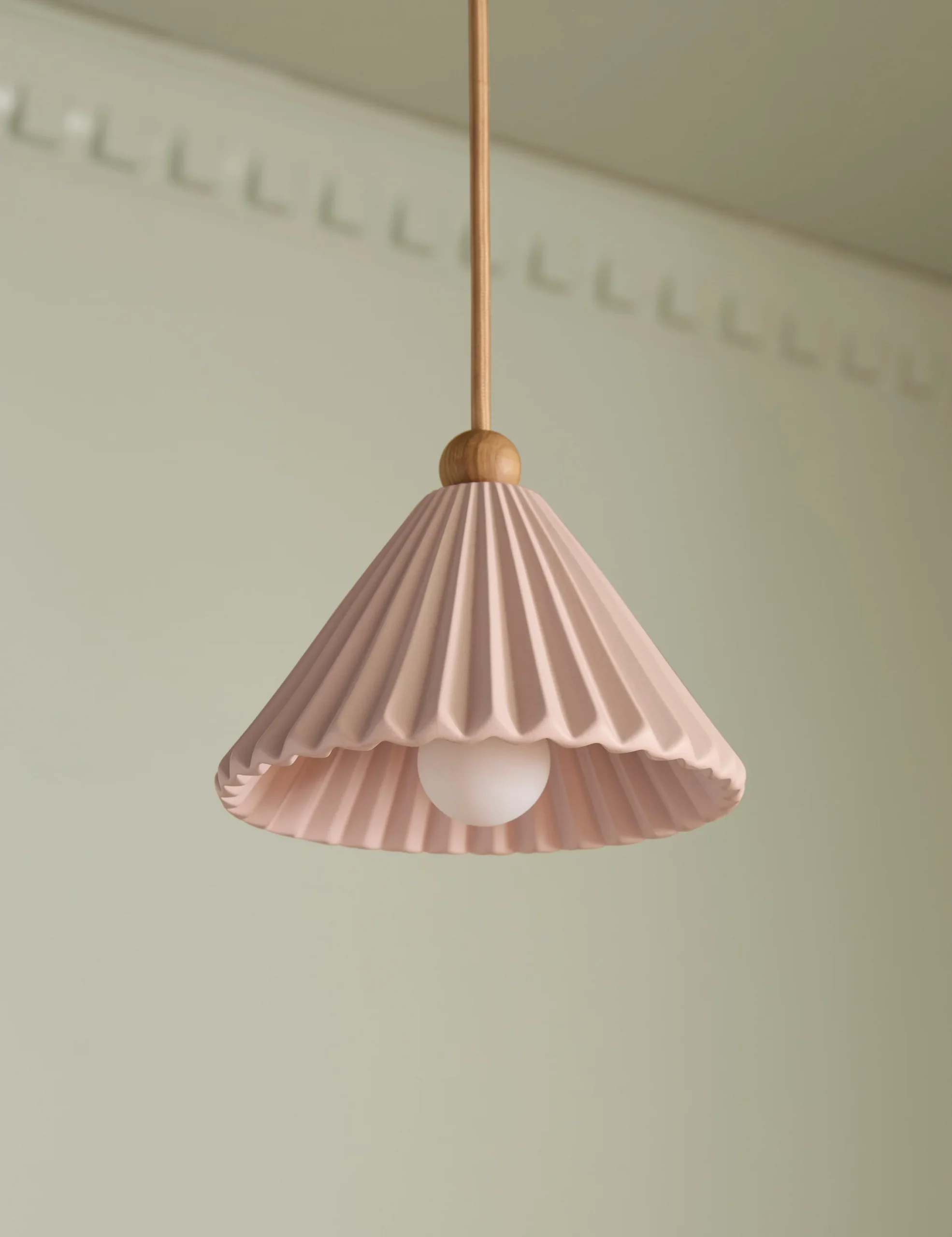 A pink pleated pendant lampshade with a wooden fitting hangs from the ceiling against a light-colored wall.
