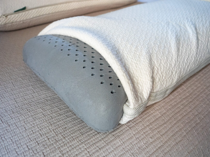 A perforated gray foam pillow is partially covered by a textured white pillowcase and placed on a beige bedspread.