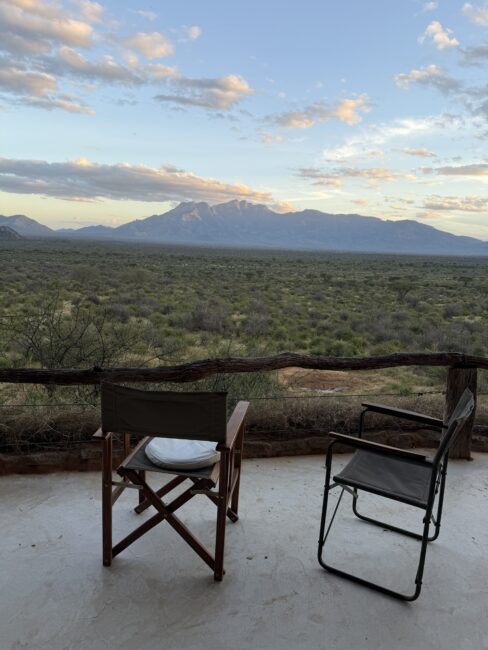 Two empty chairs on a patio overlook a vast green landscape with distant mountains under a partly cloudy sky at sunset.