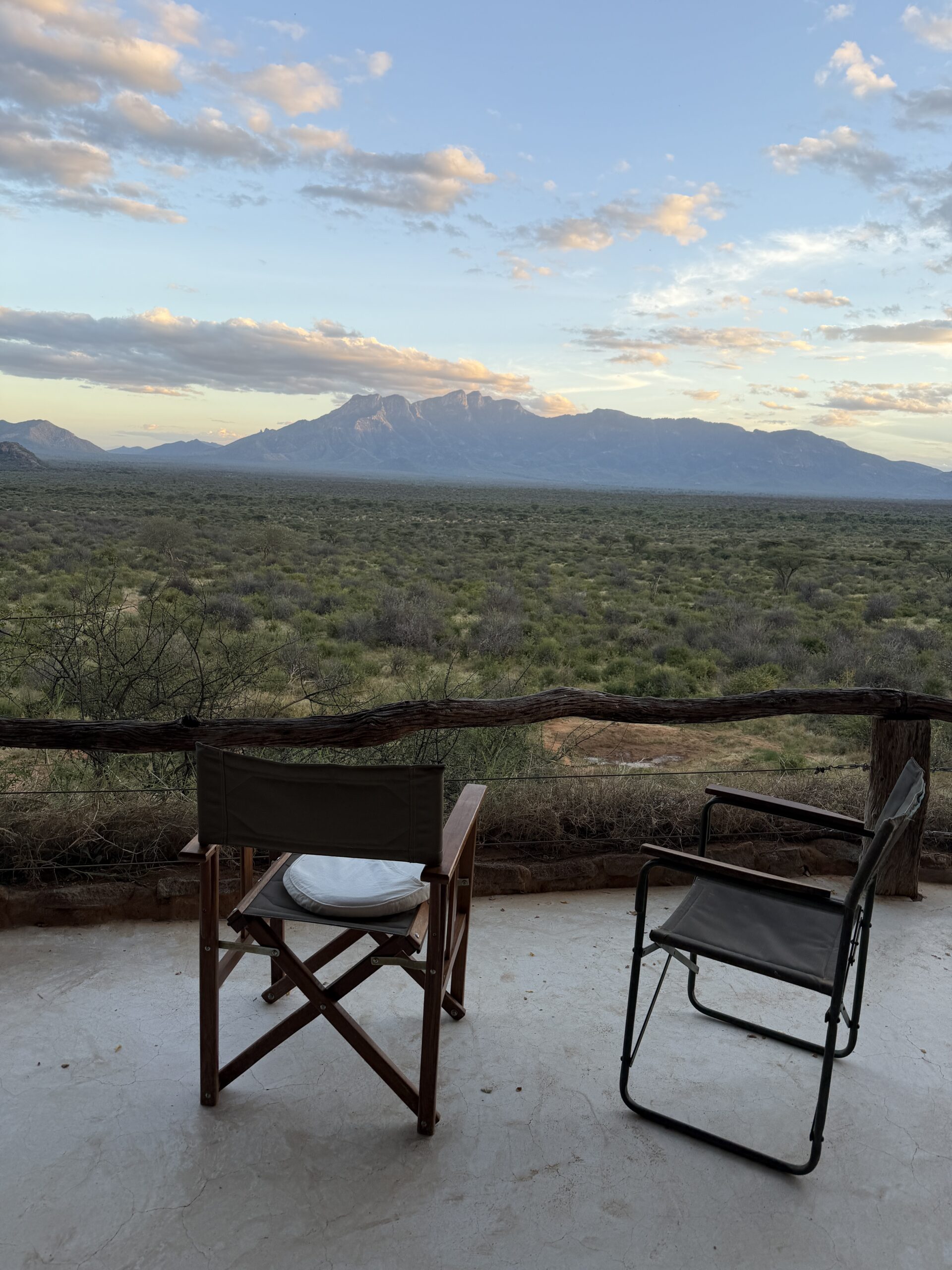 Two empty chairs on a patio overlook a vast green landscape with distant mountains under a partly cloudy sky at sunset.