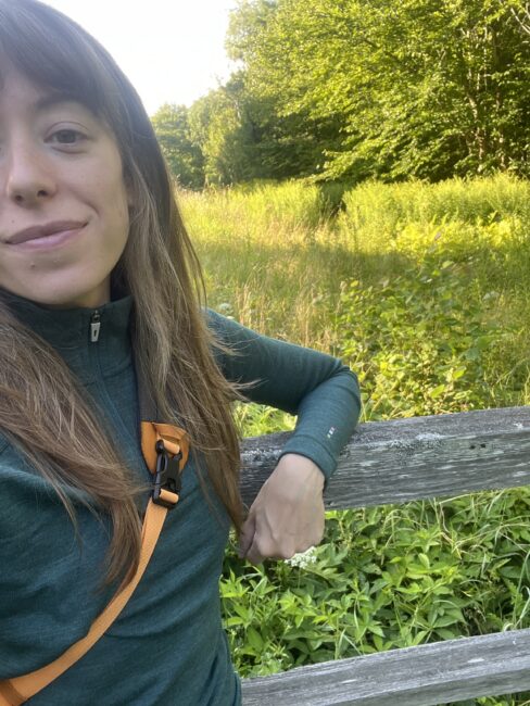 Person in a green jacket stands by a wooden fence in a lush, sunlit field with tall grass and trees in the background.