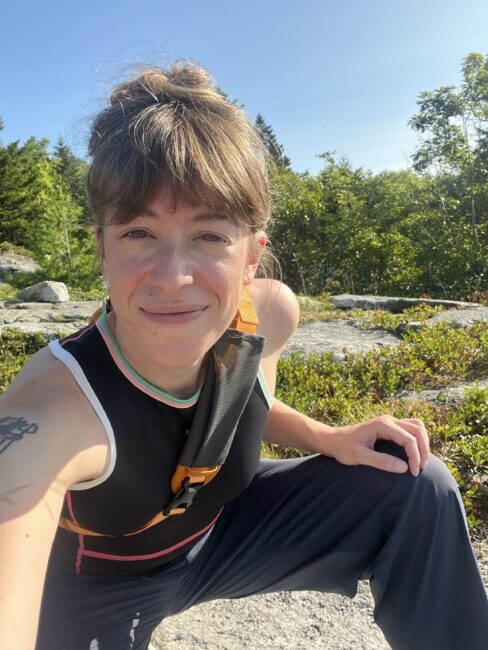 Person in athletic clothing takes a selfie outdoors on a sunny day, surrounded by greenery and rocks.