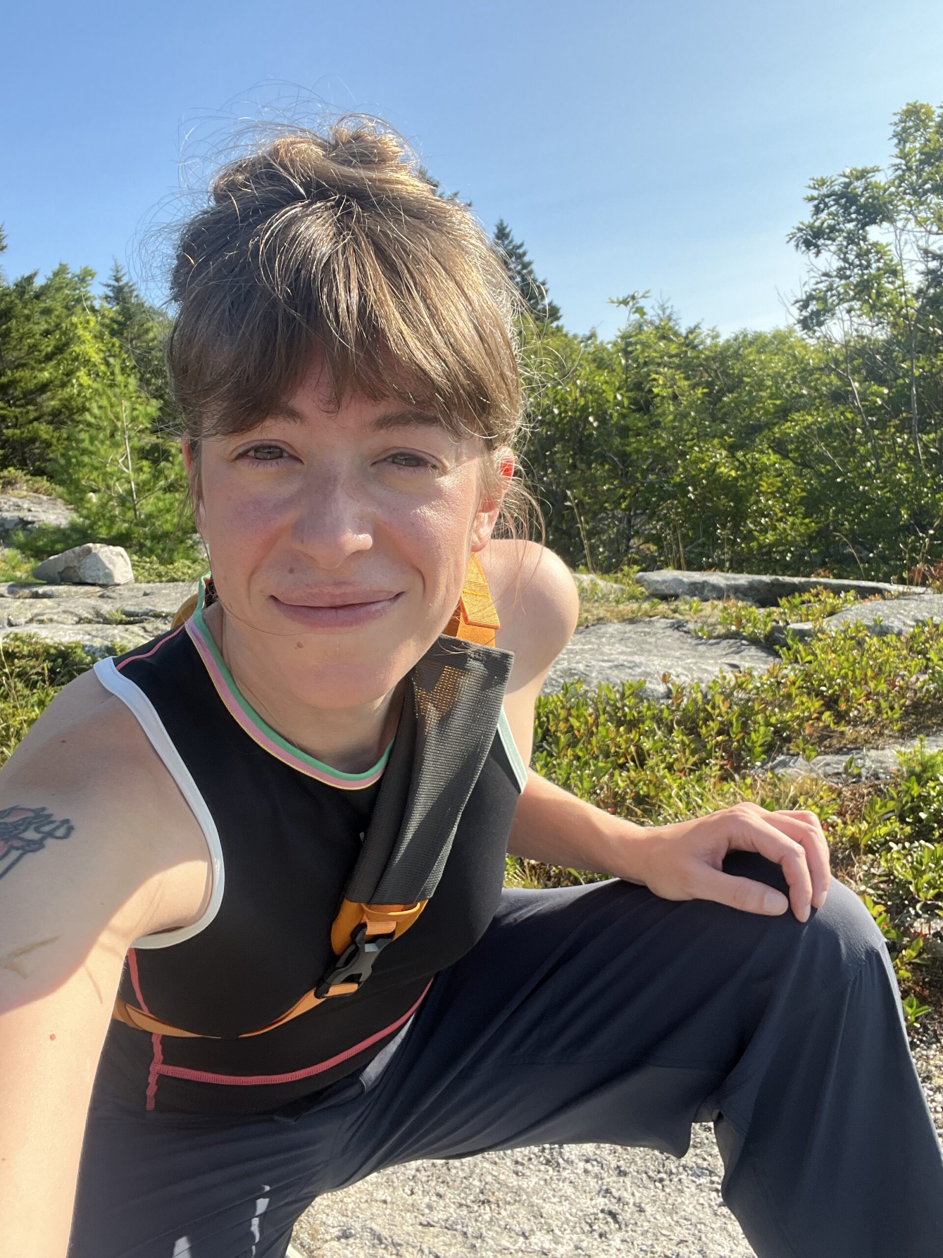 Person in athletic clothing takes a selfie outdoors on a sunny day, surrounded by greenery and rocks.