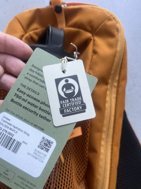 A hand holds green and white product tags attached to an orange backpack, featuring a “Fair Trade Certified Factory” label.