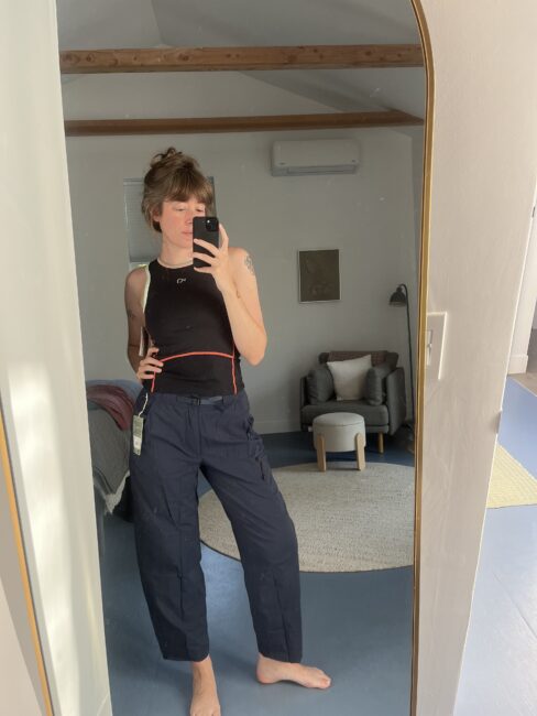 Person taking a mirror selfie in a bedroom, wearing a black tank top and dark cargo pants with a tag attached. A modern chair, lamp, and rug are visible in the background.