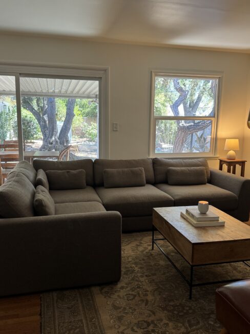 A gray sectional sofa sits in a living room with a wooden coffee table, a rug, and large windows looking out to a green backyard.