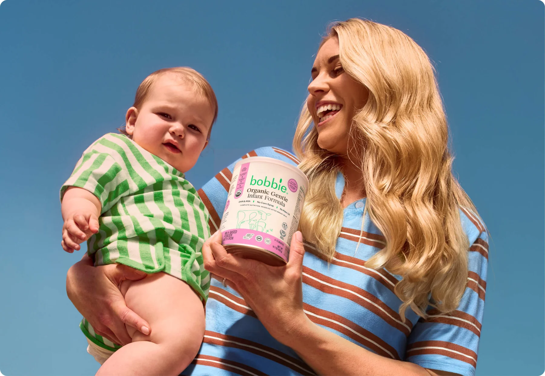 A woman holding a baby in one arm and a container of Bobbie organic infant formula in the other, both outdoors under a clear blue sky.