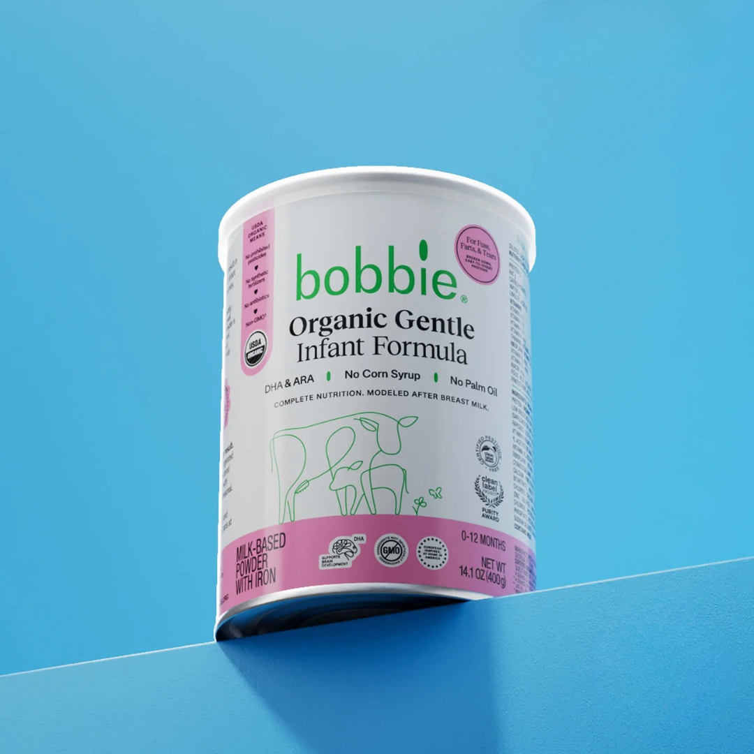 A can of Bobbie Organic Gentle Infant Formula with a white and pink label is displayed on a blue surface against a blue background.