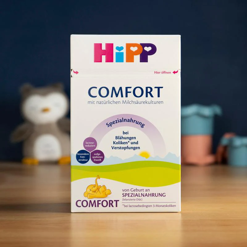A box of HiPP Comfort infant formula stands on a wooden surface, with blurred nursery items in the background.