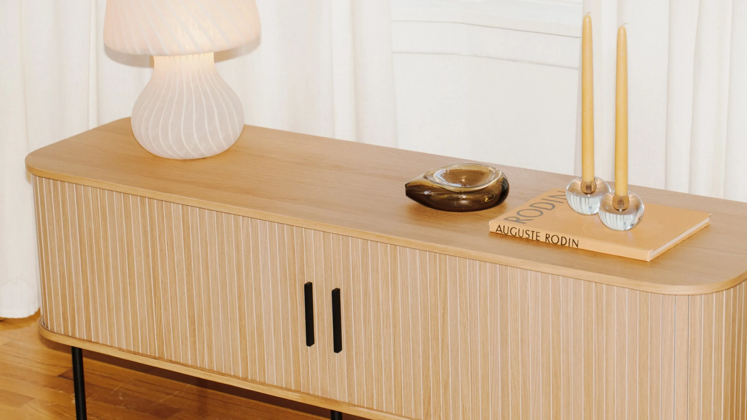 A light wood sideboard with vertical grooves holds a white lamp, a glass dish, a book titled “Rodin,” and two tall yellow candles on a tray.