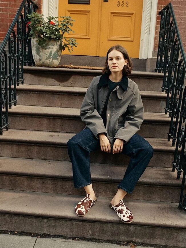 A person sits on brownstone steps near a large planter, wearing a gray jacket, dark pants, and cow-print shoes. A yellow door with the number 332 is in the background.