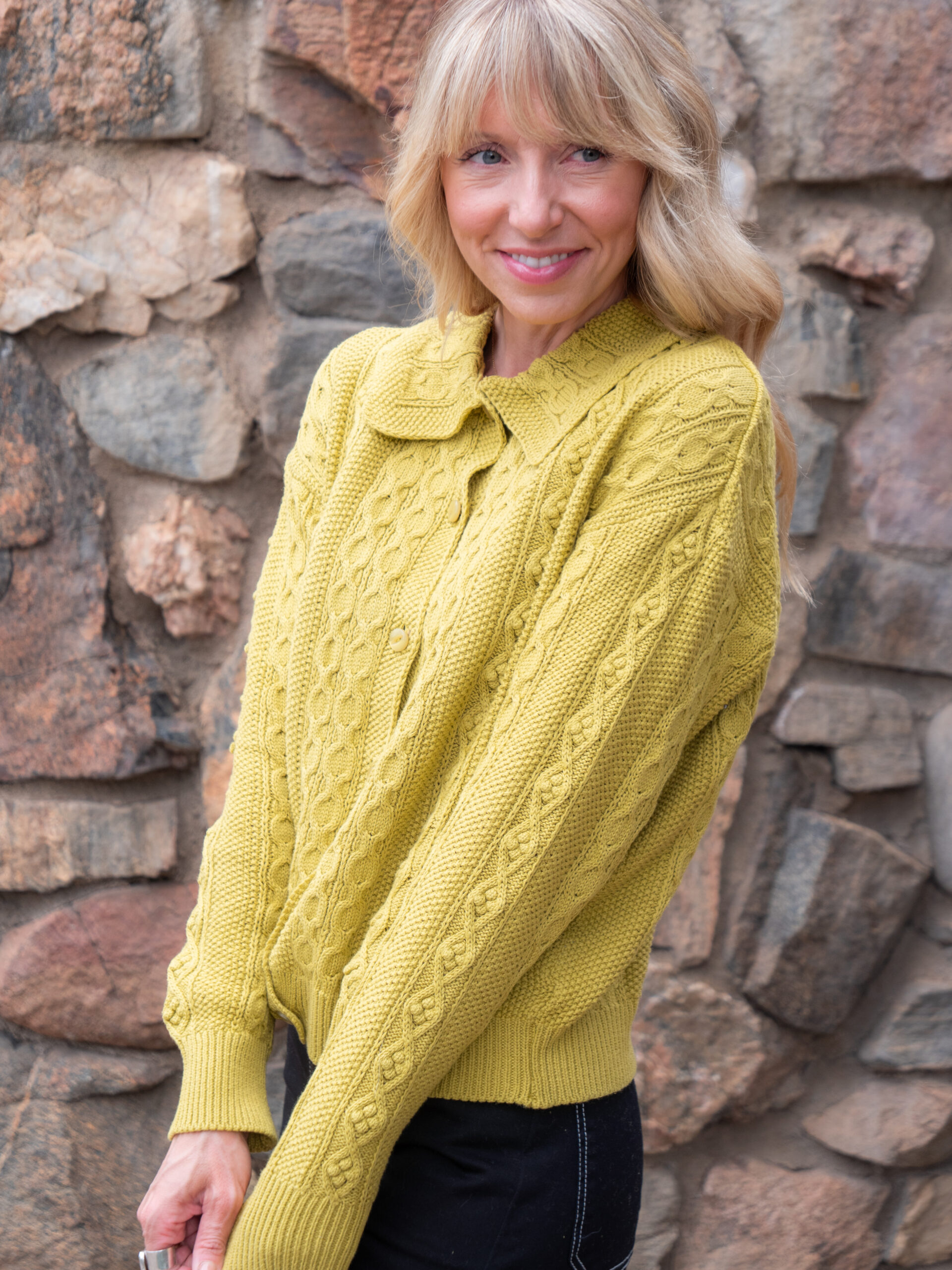 A woman wearing a yellow cable-knit sweater stands in front of a stone wall, smiling slightly with her arms relaxed in front of her.