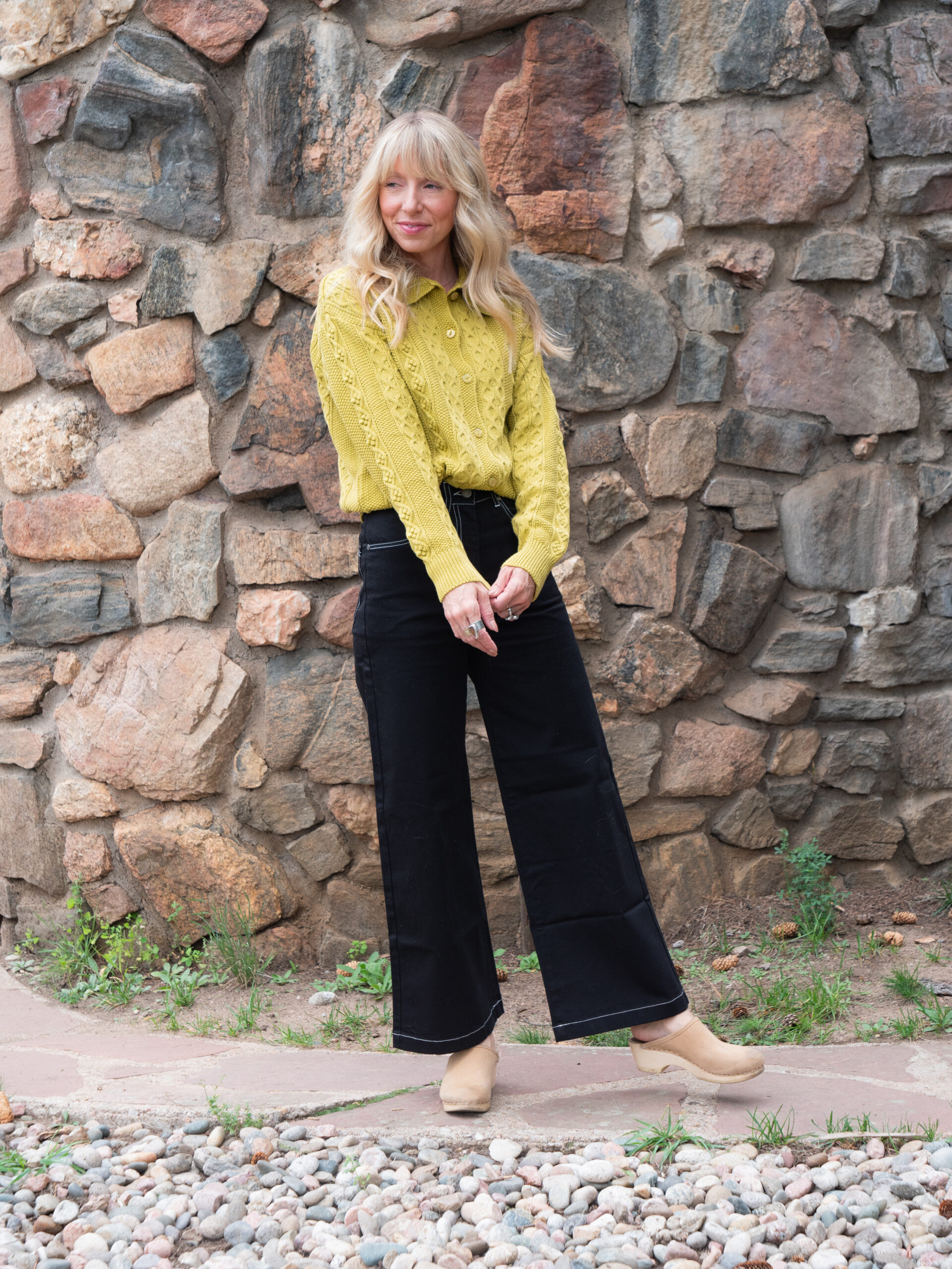 A woman stands on a stone path in front of a rock wall, wearing a yellow blouse, black wide-leg pants, and beige shoes.