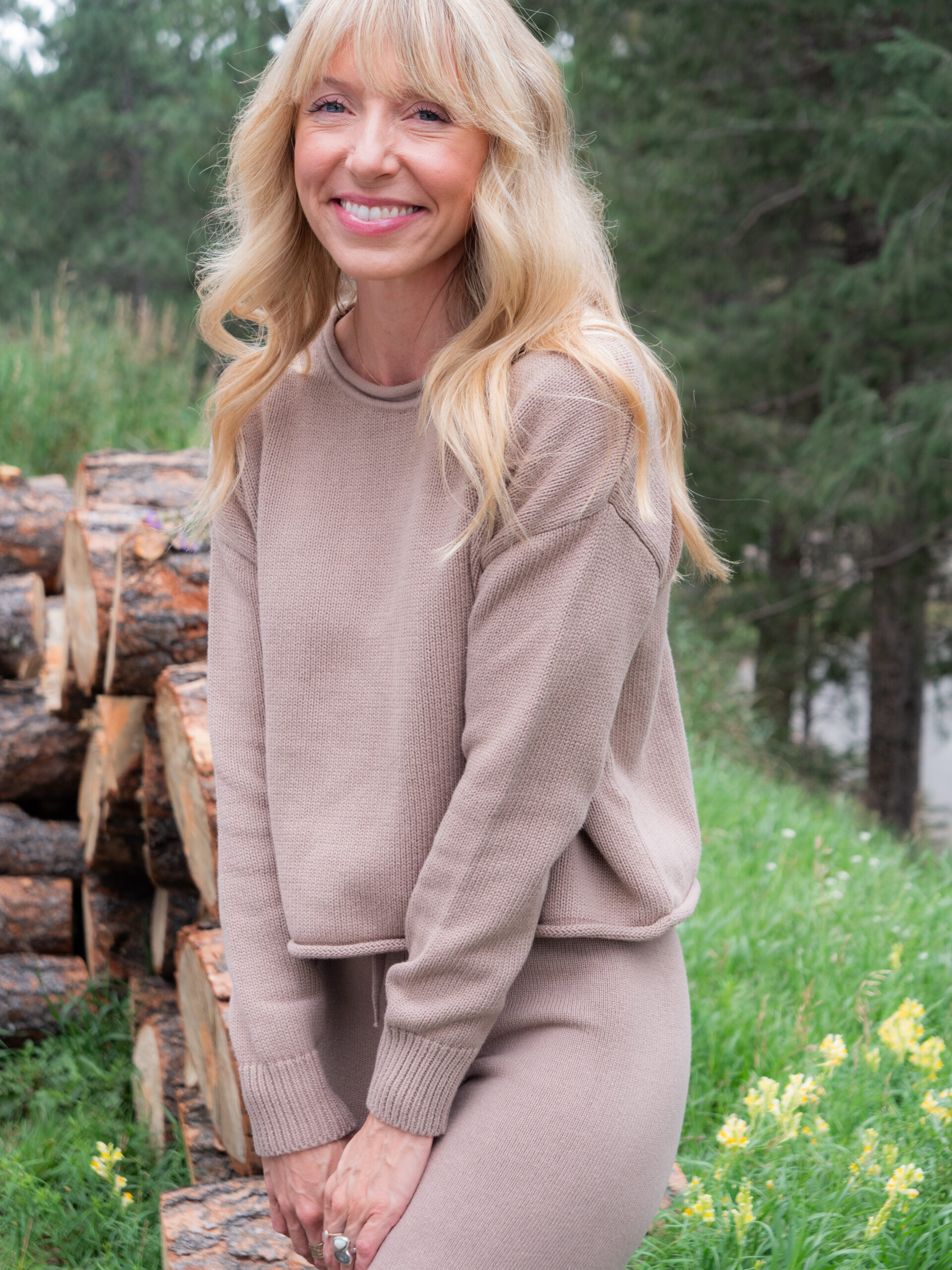 Woman with long blonde hair in a beige sweater and pants stands outdoors near stacked logs and green grass, smiling at the camera.
