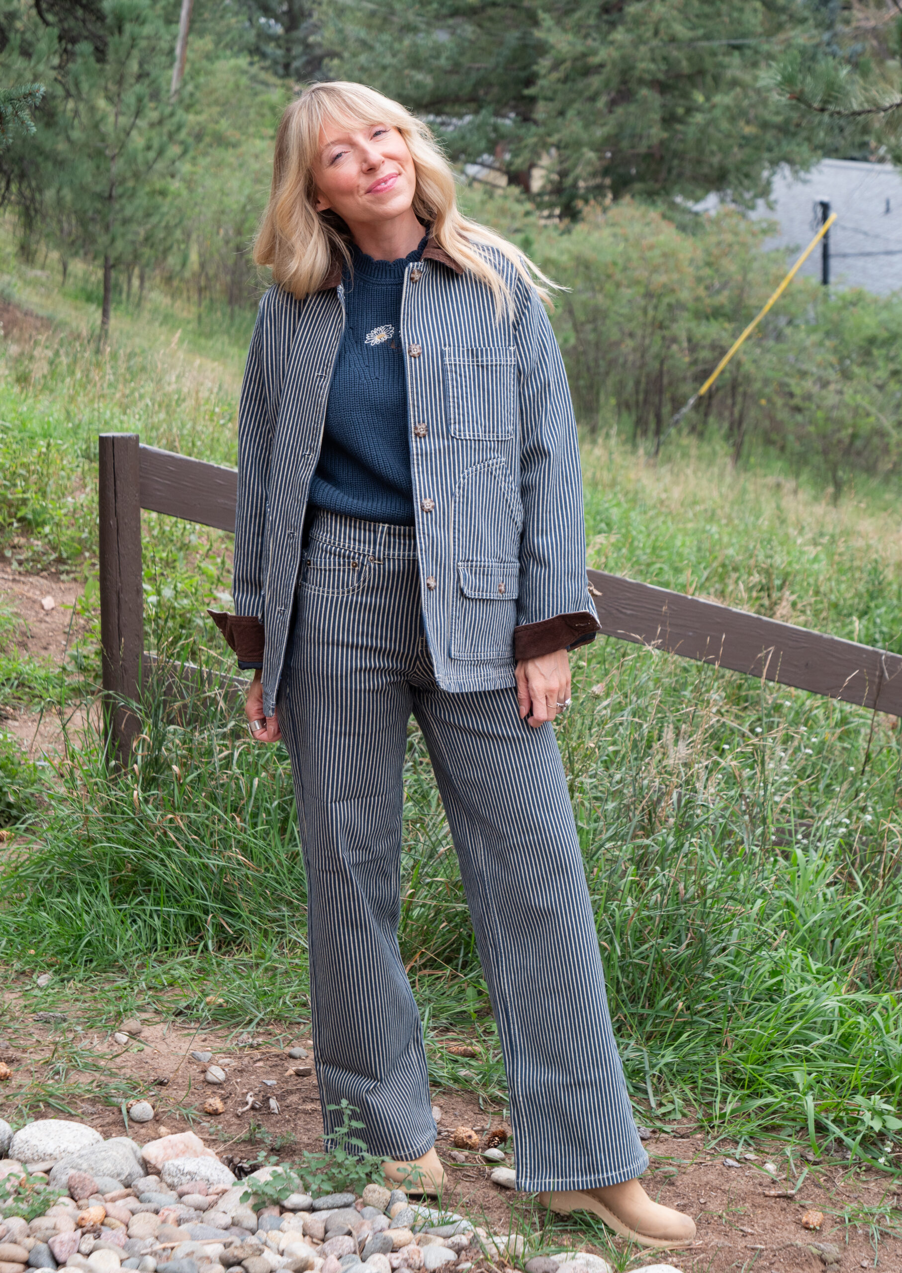 Woman stands outdoors on a rocky path, wearing a blue striped jacket and matching pants over a blue sweater, with greenery and trees in the background.