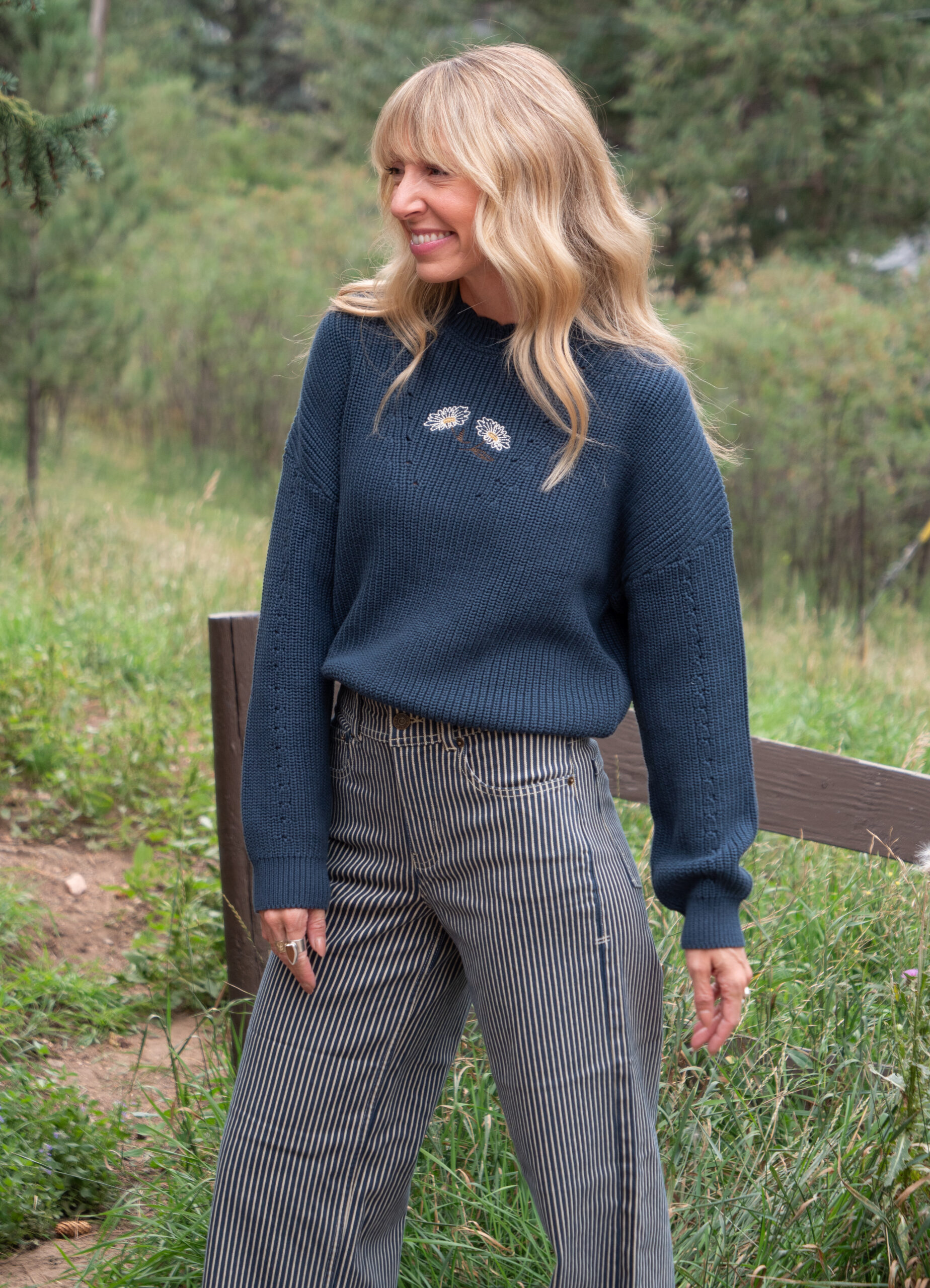 Woman with long blonde hair wearing a dark blue sweater and striped pants stands outdoors, smiling with greenery in the background.