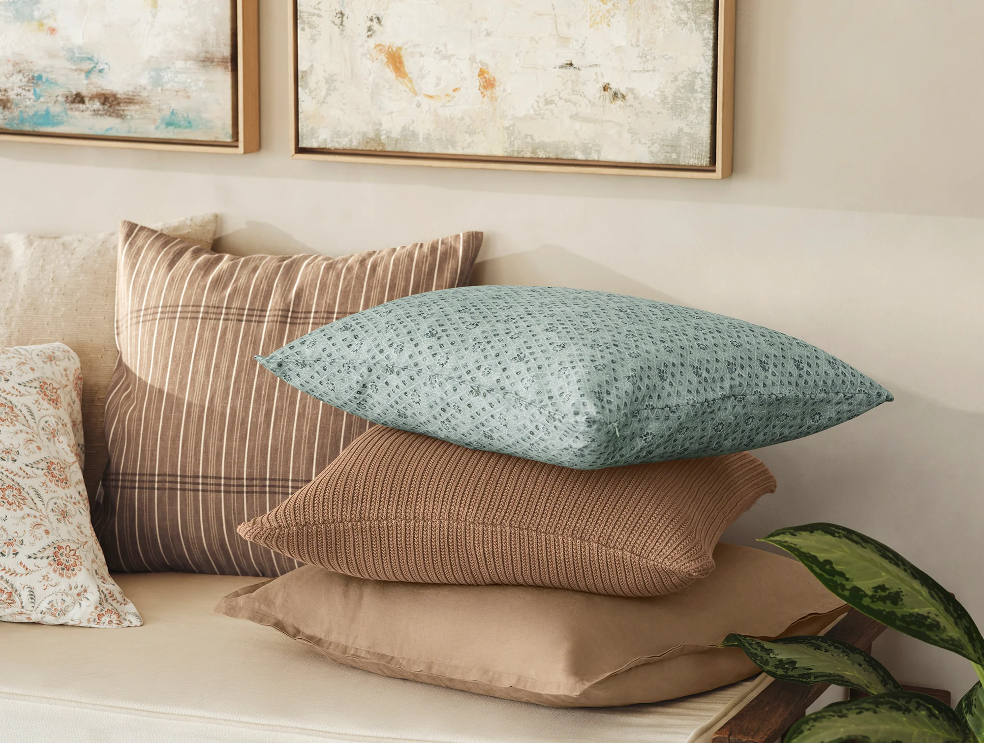Four patterned and solid pillows are stacked on a beige sofa with a green leafy plant in the foreground and abstract artwork hanging on the wall above.