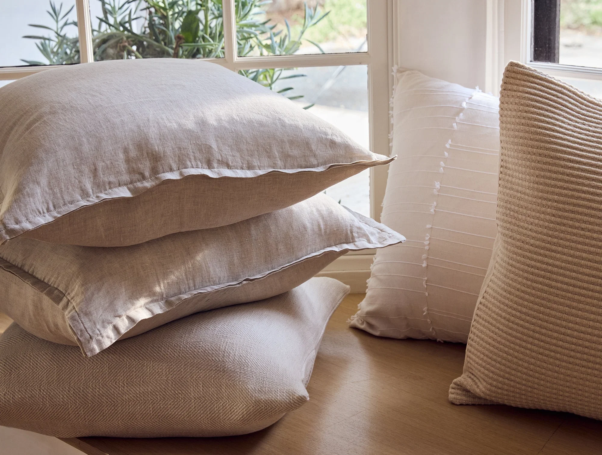 Four beige and off-white pillows are stacked and placed on a wooden surface near a window with plants visible outside.