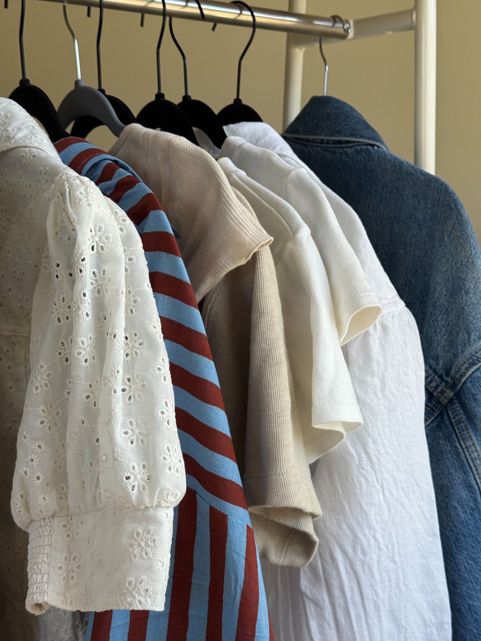 A clothing rack holding assorted garments, including a white eyelet blouse, a striped shirt, neutral tops, a white shirt, and a blue denim jacket, all on black hangers.