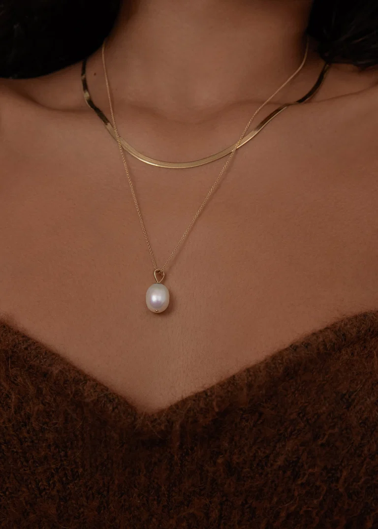 A person wears a gold chain with a flat design and a second gold necklace featuring a single pearl pendant, paired with a brown textured top.