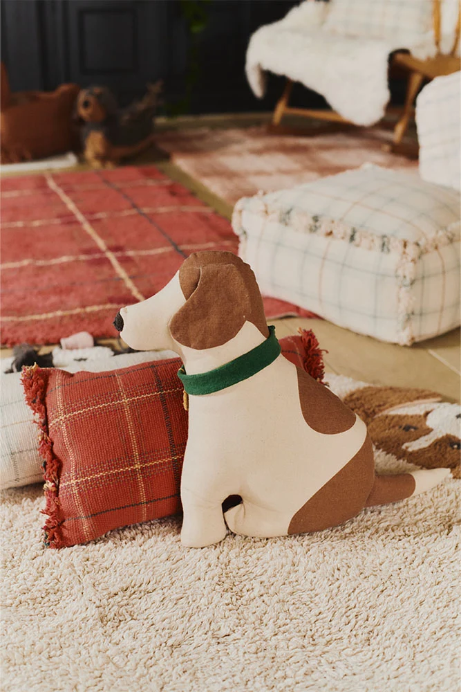 A plush dog-shaped doorstop with brown patches sits on a beige rug next to a red plaid pillow in a cozy, carpeted room.