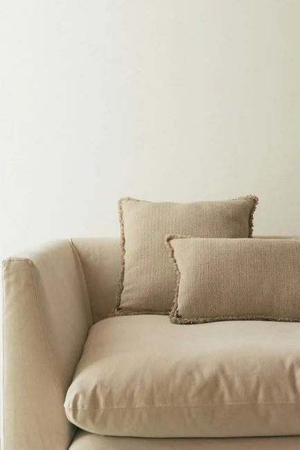 A beige couch with two textured beige pillows sits against a plain off-white wall.