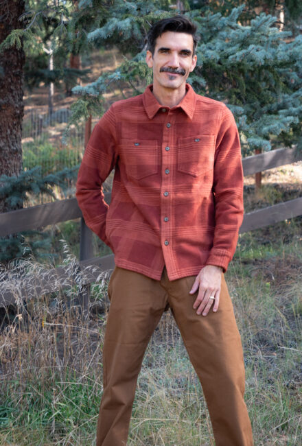 A man with a mustache wears a rust-colored button-up shirt and brown trousers, standing in a wooded area.