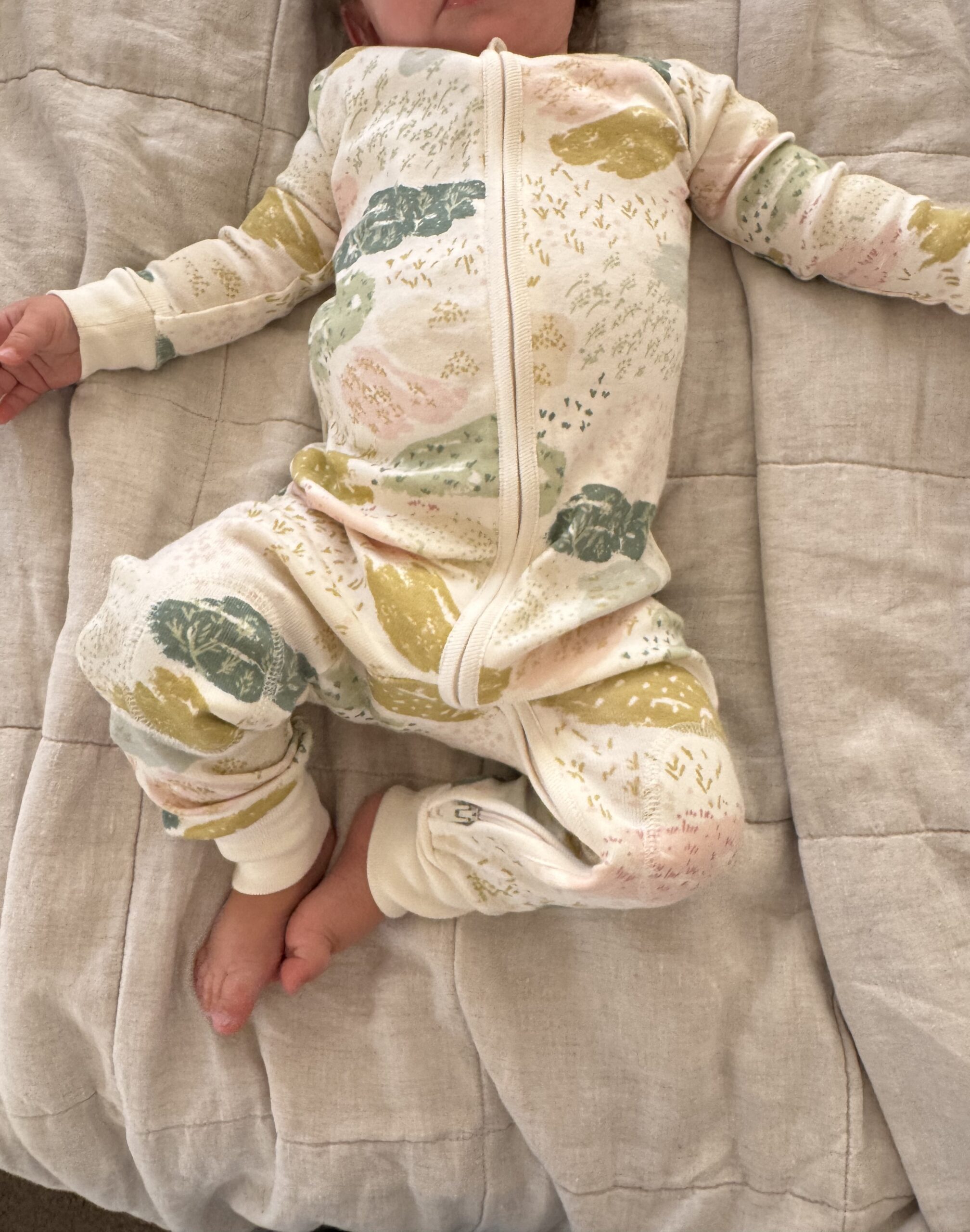 Baby in a cream-colored, patterned onesie lies on a beige quilted blanket, with arms and legs relaxed and face partially out of frame.